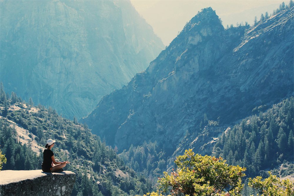 Person meditating on cliff overlooking mountainous landscape.